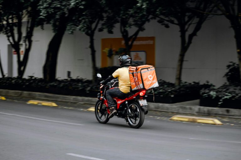 A delivery man riding a motorcycle with a thermal bag on a city street during the day.