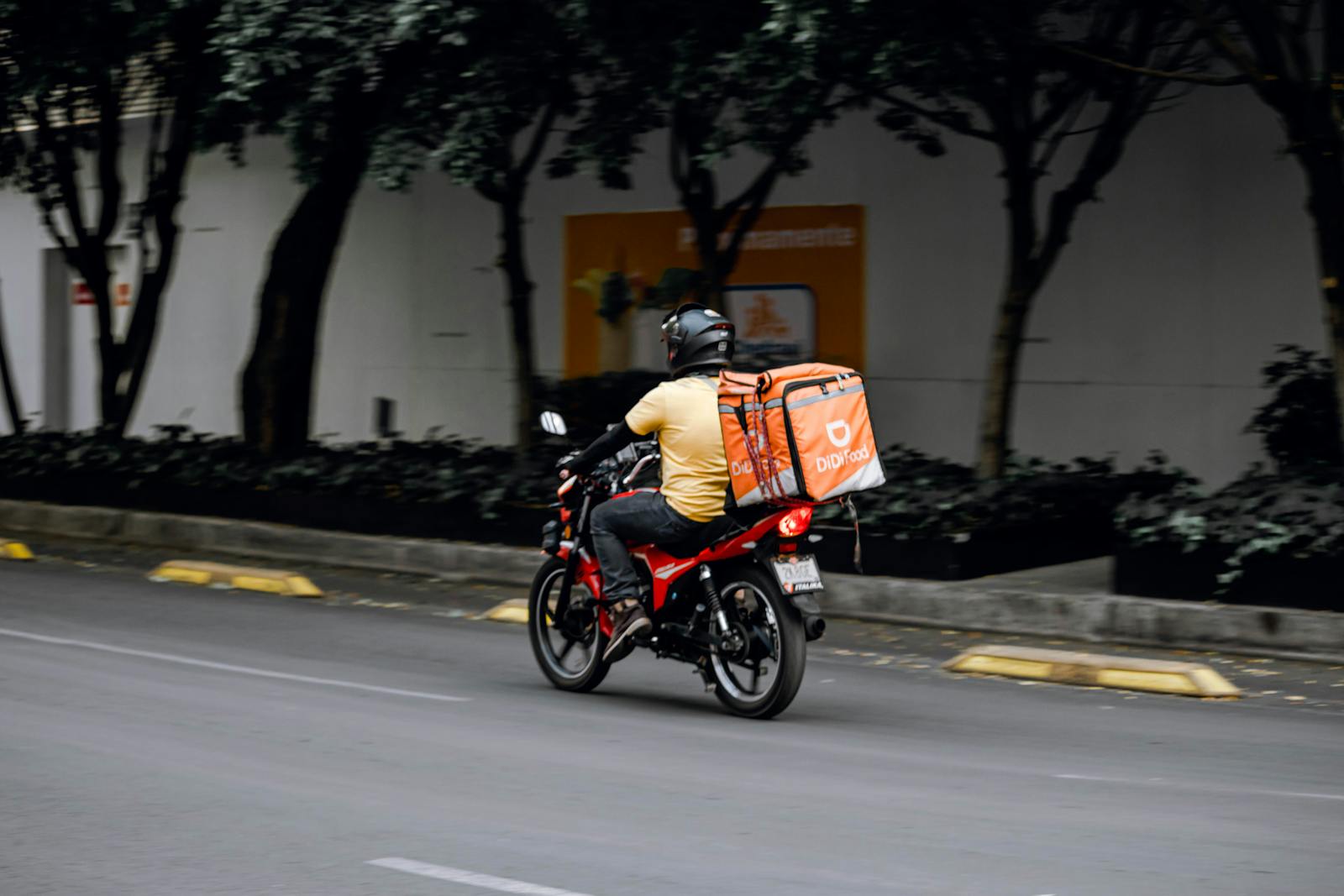 A delivery man riding a motorcycle with a thermal bag on a city street during the day.